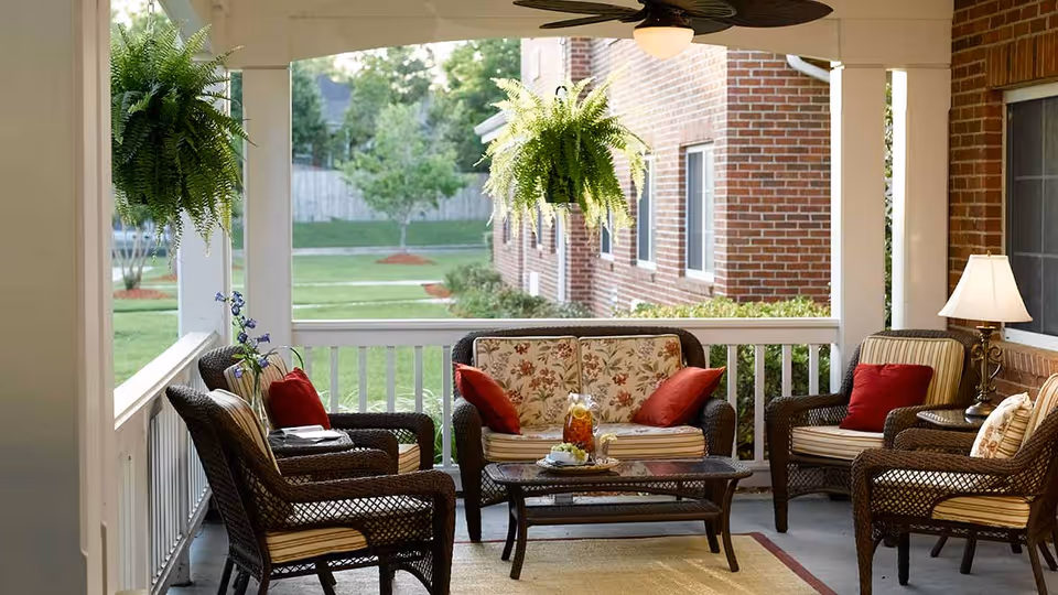 Covered outdoor patio area with wicker furniture including a loveseat and four chairs with cushions and red pillows, a glass coffee table with a pitcher of iced tea and glasses, two hanging green ferns, a ceiling fan, and a table lamp. The patio overlooks a grassy yard with trees and a brick building.