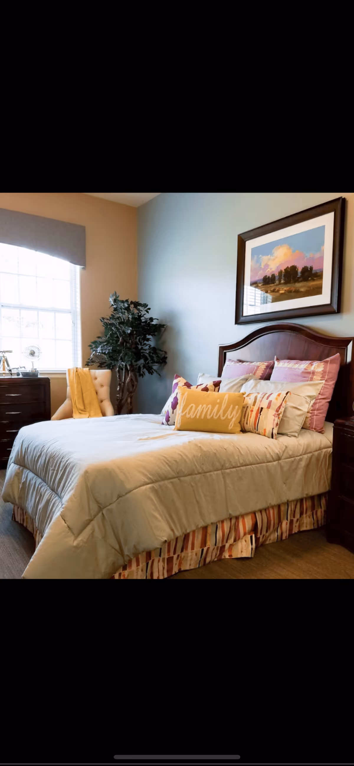 A neatly made bedroom with a bed topped with decorative pillows (one reading 'family'), a wooden headboard, framed artwork above, a potted plant, dresser and a window.