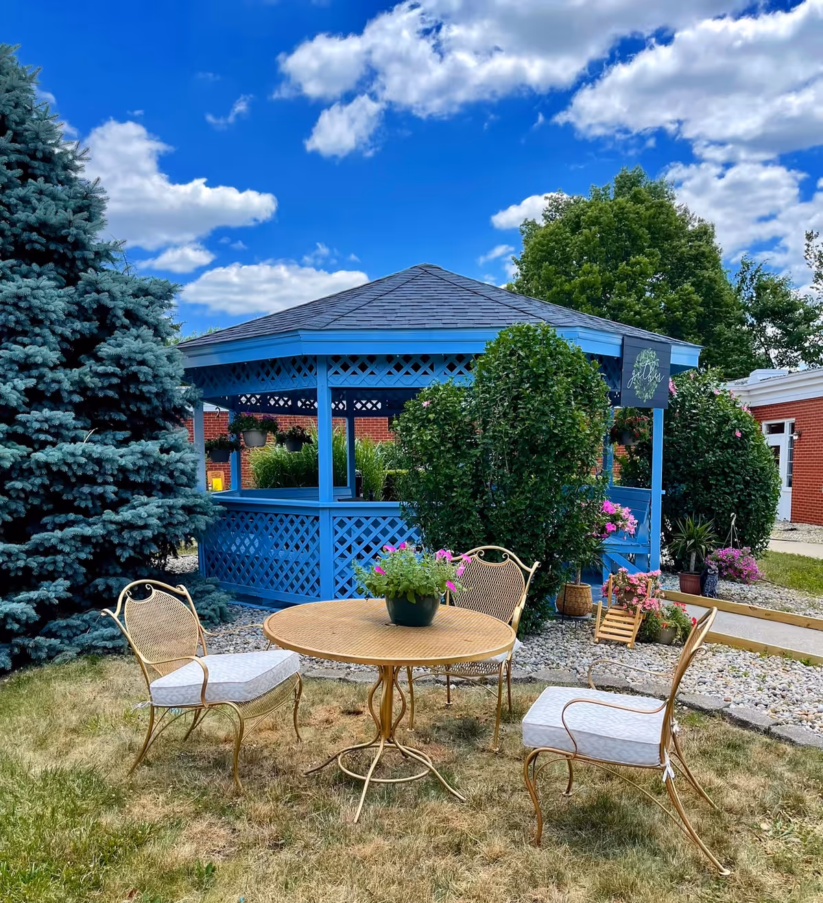 Blue wooden gazebo surrounded by shrubs and flowers with a round metal table and three cushioned chairs on a grassy lawn under a blue sky.