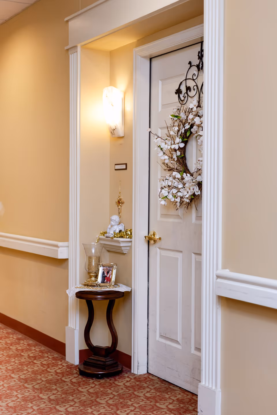 A hallway corner in an assisted living facility with beige walls and patterned carpet. A white door decorated with a floral wreath is framed by white molding. Next to the door is a small wooden table holding a glass candle holder and a framed photo. Above the table is a small wall shelf with a white angel figurine and gold decorations. A wall sconce light fixture illuminates the area.