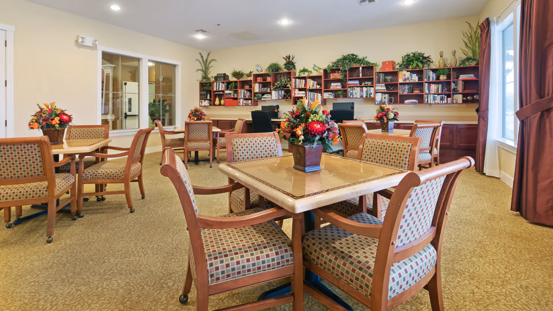 A well-lit room with multiple square tables surrounded by wooden chairs with patterned upholstery. Each table has a floral centerpiece. The back wall features wooden shelves filled with books, plants, and decorative items. There are two computer workstations with black chairs beneath the shelves. Large windows with maroon curtains allow natural light into the room.