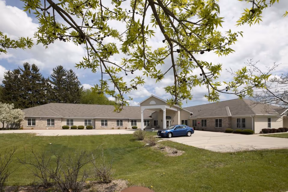 Exterior view of a single-story assisted living facility building with a covered entrance, surrounded by green grass and trees under a partly cloudy sky. A blue car is parked in front of the entrance.