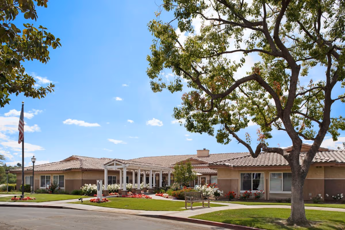 Exterior view of a single-story senior living facility building with a tiled roof, surrounded by well-maintained landscaping including green grass, flowering plants, and a large tree. There is a wooden bench on the lawn and an American flag on a flagpole near the entrance under a clear blue sky.