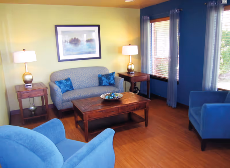 A cozy living room with a blue patterned loveseat, two blue armchairs, a wooden coffee table with decorative items, two wooden side tables each with a lamp, and a framed painting on a light yellow wall. The room has wooden flooring and two windows with blue curtains.