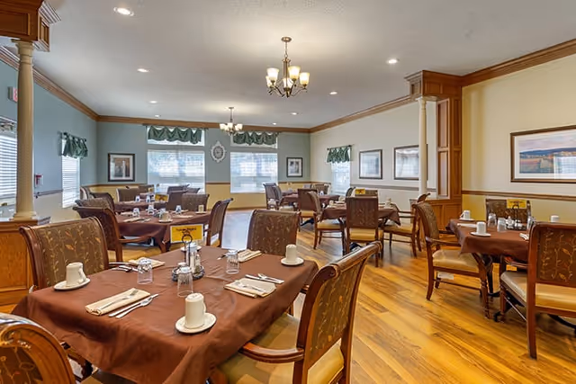A dining room with multiple tables covered in brown tablecloths, each set with white coffee cups, napkins, and silverware. The room has wooden flooring, light-colored walls with framed artwork, and windows with green valances. There are chandeliers hanging from the ceiling providing warm lighting.