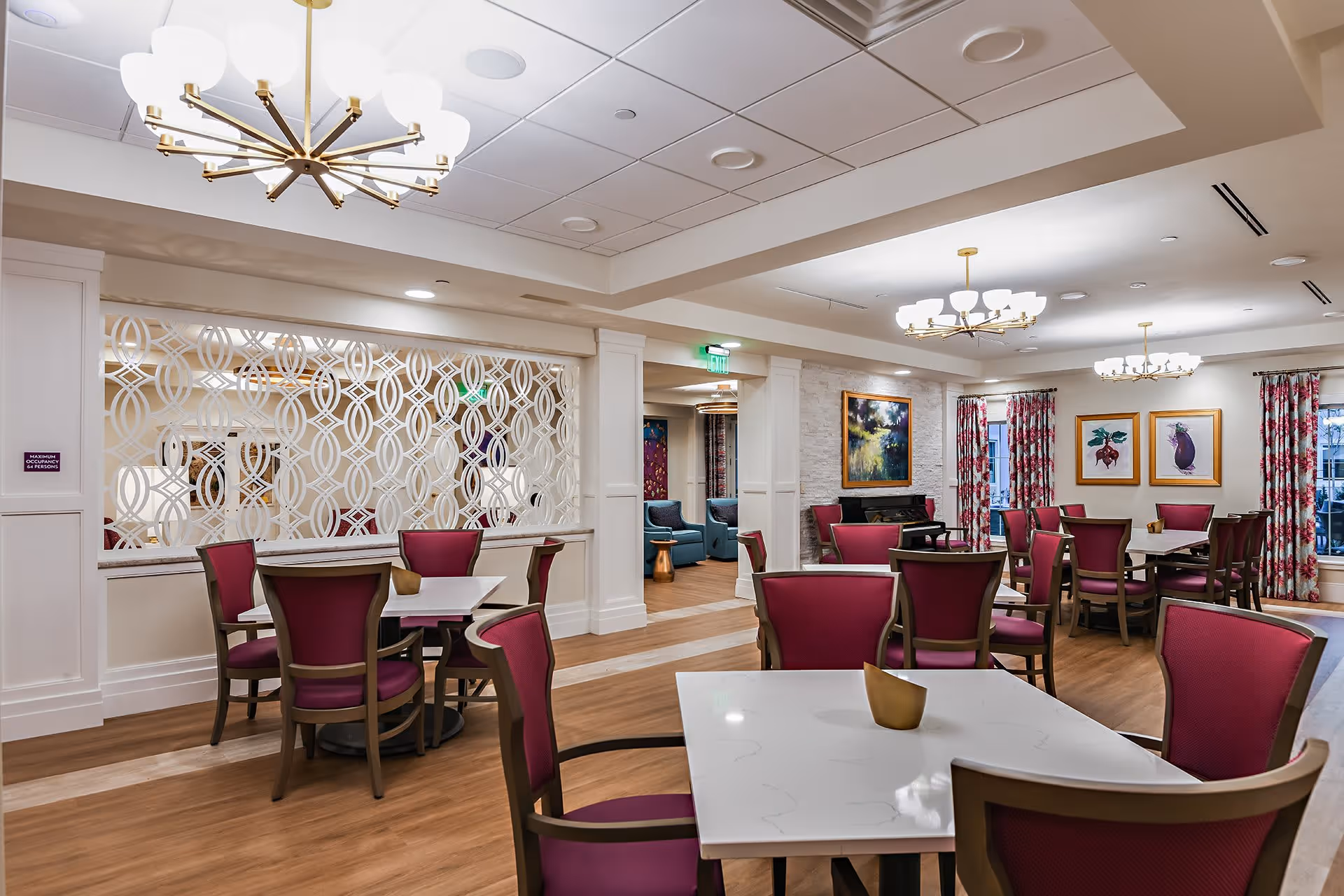 A dining room in a senior living facility with multiple tables and chairs featuring red upholstery. The room has modern chandeliers, decorative wall art, patterned curtains, and a partition with a geometric design. The flooring is wood, and there is a cozy seating area visible in the background.