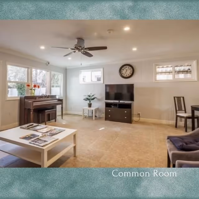 A spacious common room with a ceiling fan, a piano with a bench, a white coffee table with magazines, a flat-screen TV on a dark TV stand, a small side table with a plant, and a chair next to a round table. The room has several windows allowing natural light to enter.