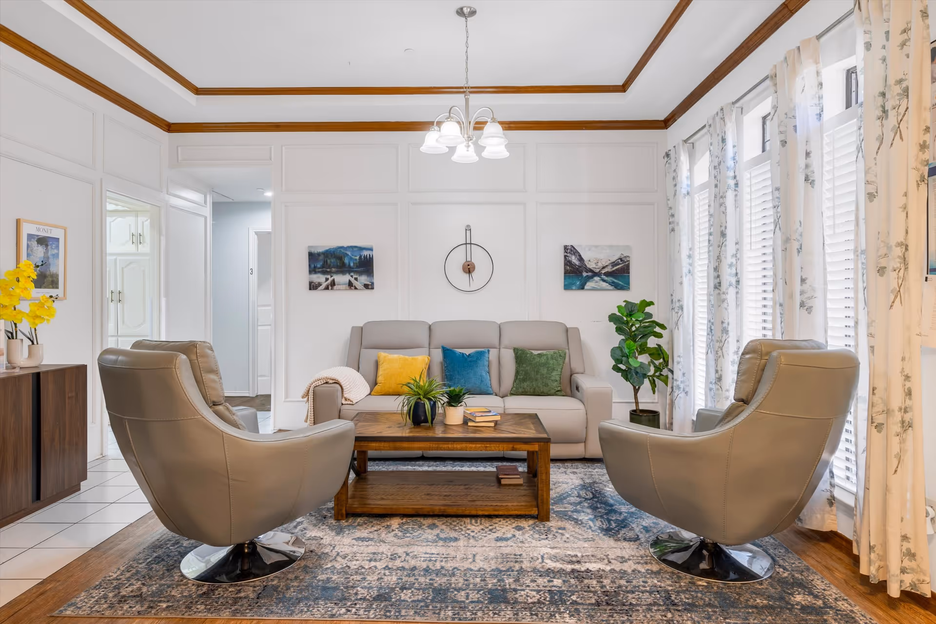 A cozy living room with a beige sofa adorned with colorful cushions in yellow, blue, and green. Two modern beige swivel chairs face the sofa across a wooden coffee table with plants and books. The room has white walls with wooden trim, a patterned area rug, and large windows with floral curtains allowing natural light to fill the space. Wall art and a decorative clock hang above the sofa.