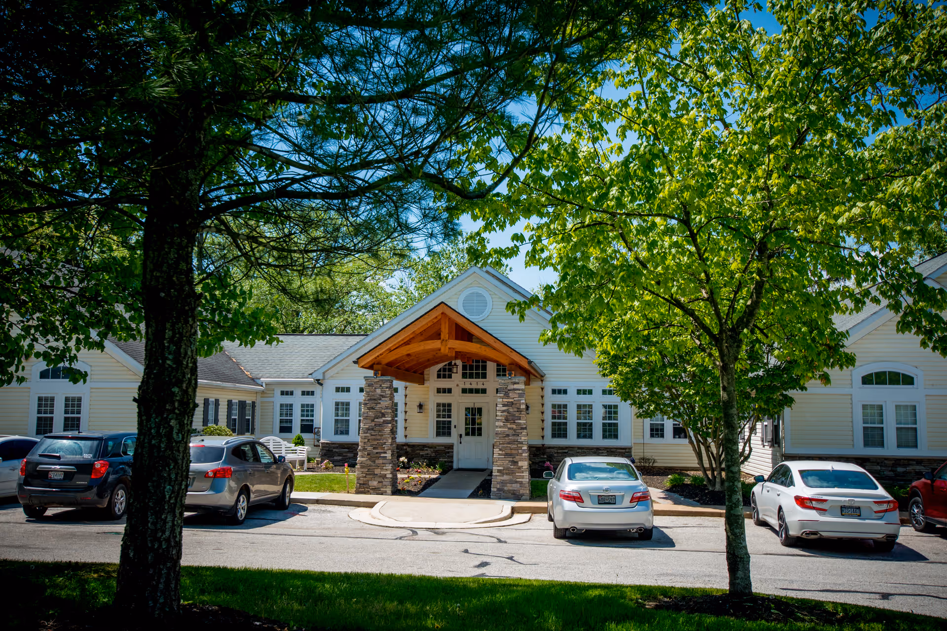 Front exterior view of Inspirations Memory Care of Lutherville building with a covered entrance supported by stone pillars, surrounded by trees and parked cars in the driveway.