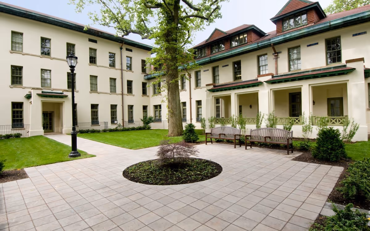 Outdoor courtyard area of a senior living facility with a tiled walkway, green grass, a large tree in the center, benches, and a multi-story building with multiple windows and a green roof in the background.