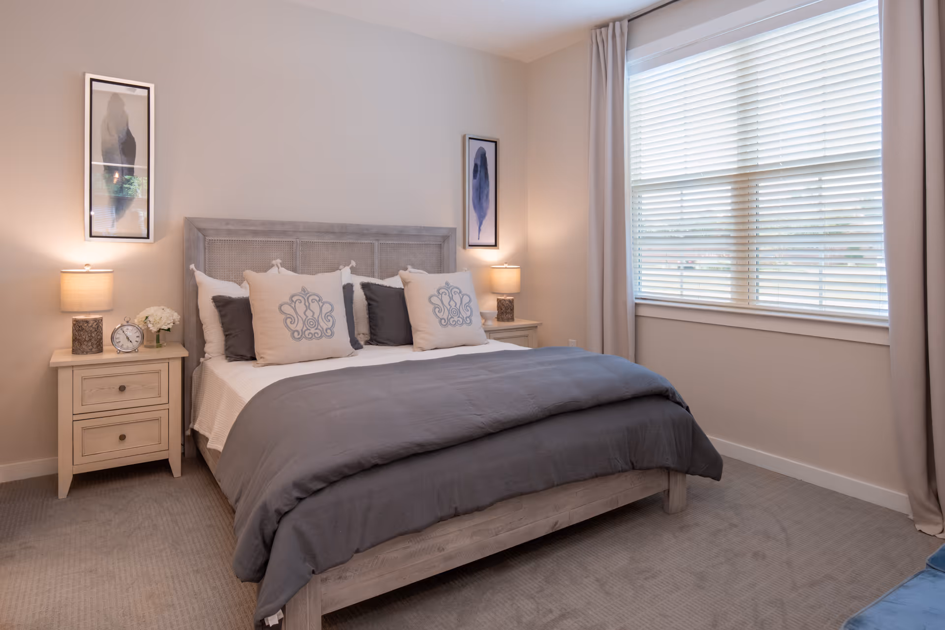 A neatly made bed with gray and beige bedding in a bedroom. The bed has a wooden headboard and is flanked by two matching nightstands, each with a lamp and decorative items. Two framed abstract art pieces hang on the wall above the nightstands. A large window with white blinds and beige curtains is on the right side of the room.