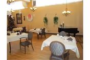 Dining room with tables covered in white tablecloths, chairs, a piano, wall plants, and a decorated Christmas tree.