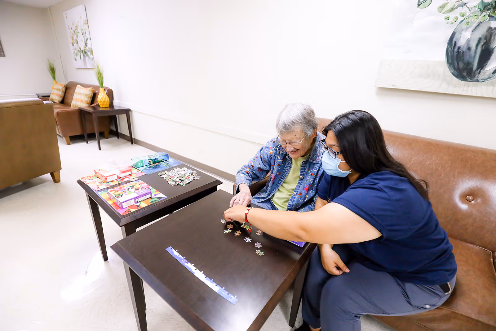 An elderly woman and a caregiver wearing a face mask are sitting on a brown couch in a well-lit room. They are working together on assembling a jigsaw puzzle on a dark wooden table in front of them. Another table nearby holds more puzzle boxes and pieces. The room has light-colored walls and floors, with additional seating and decorative plants in the background.