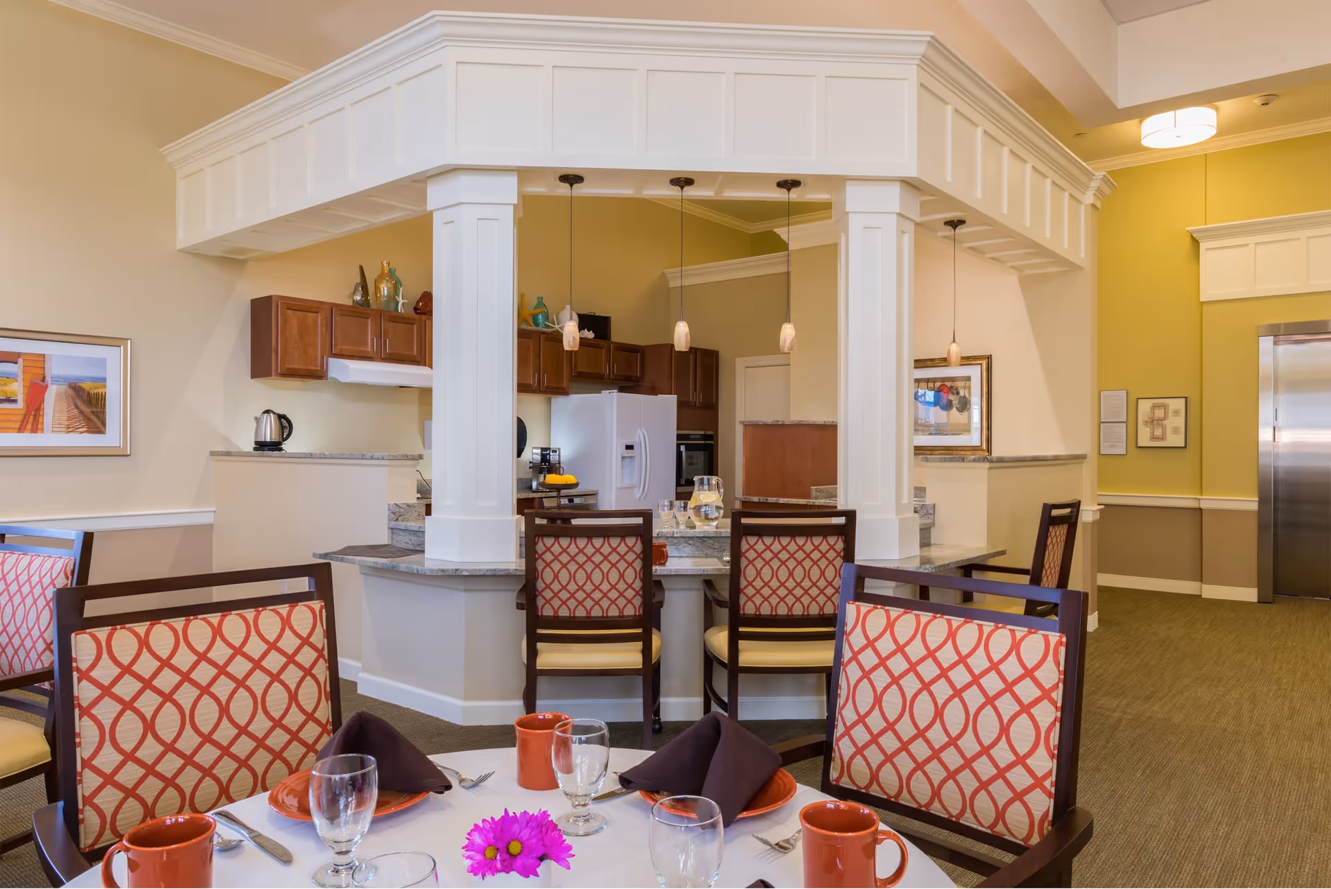Dining area with a round table set with orange plates, brown napkins, glasses, and orange mugs. The chairs have a red and beige patterned upholstery. In the background, there is a kitchen area with a white refrigerator, wooden cabinets, and a granite countertop with three pendant lights hanging above it. The walls are painted in beige and green tones, and there is framed artwork on the walls.