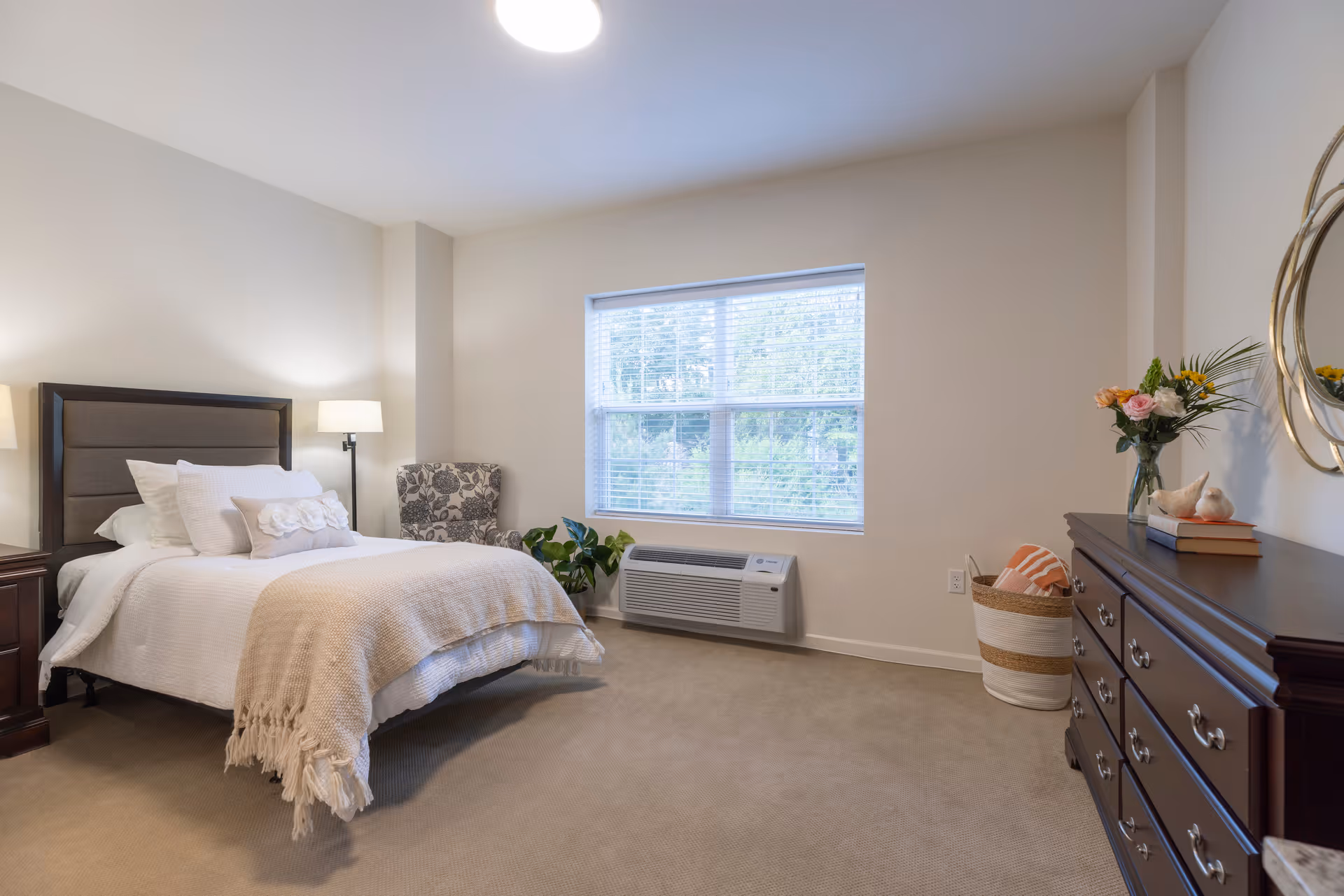 Sunlit bedroom with a made bed, armchair, dresser, and a window air-conditioning unit in a neutral-decor room.