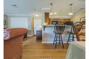 View of a senior living facility interior showing a living area with a patterned armchair, a kitchen with wooden cabinets, a breakfast bar with three black wooden bar stools, and a dining table with a checkered tablecloth. The floor is wooden, and the walls are light-colored with some framed artwork.