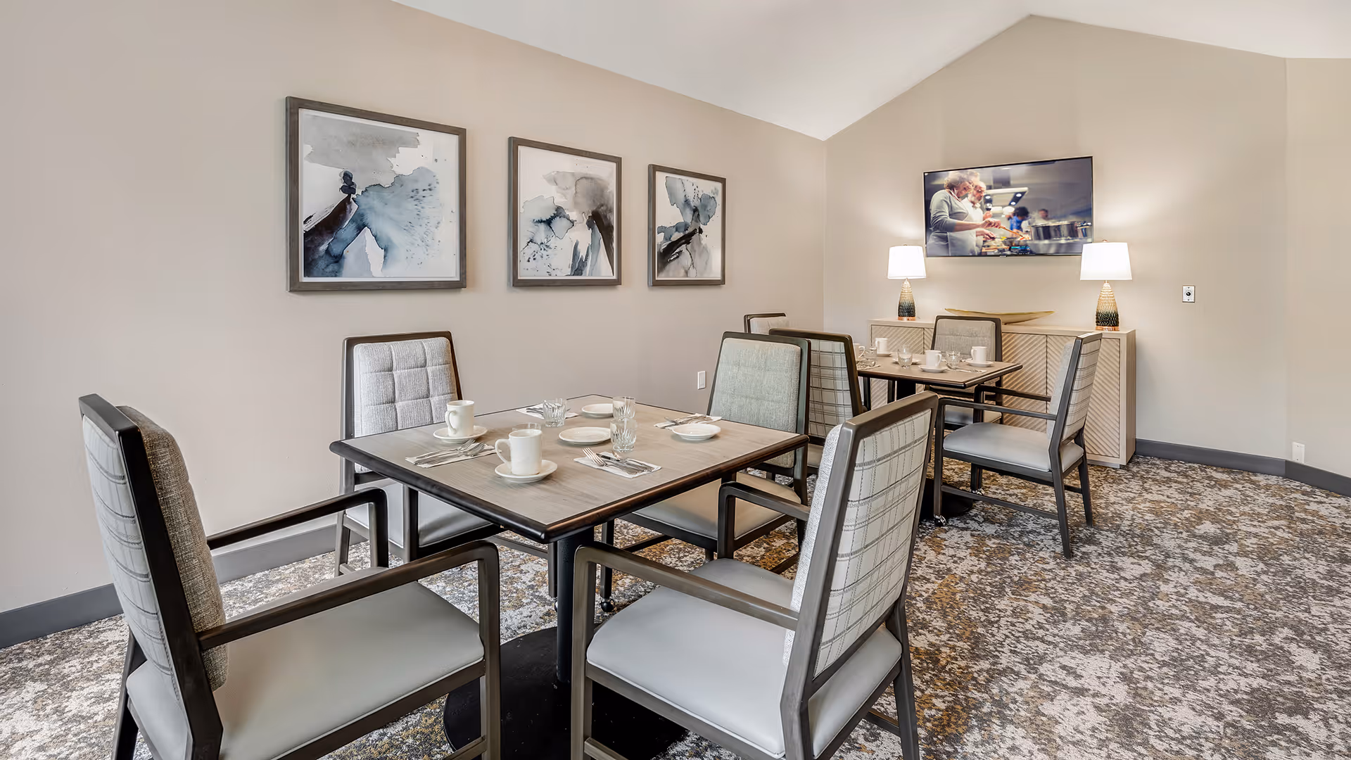 A dining room with two square tables, each set with white cups, plates, and silverware. The room features six cushioned chairs with armrests, a patterned carpet, three abstract paintings on the wall, and a flat-screen TV mounted above a sideboard with two table lamps.