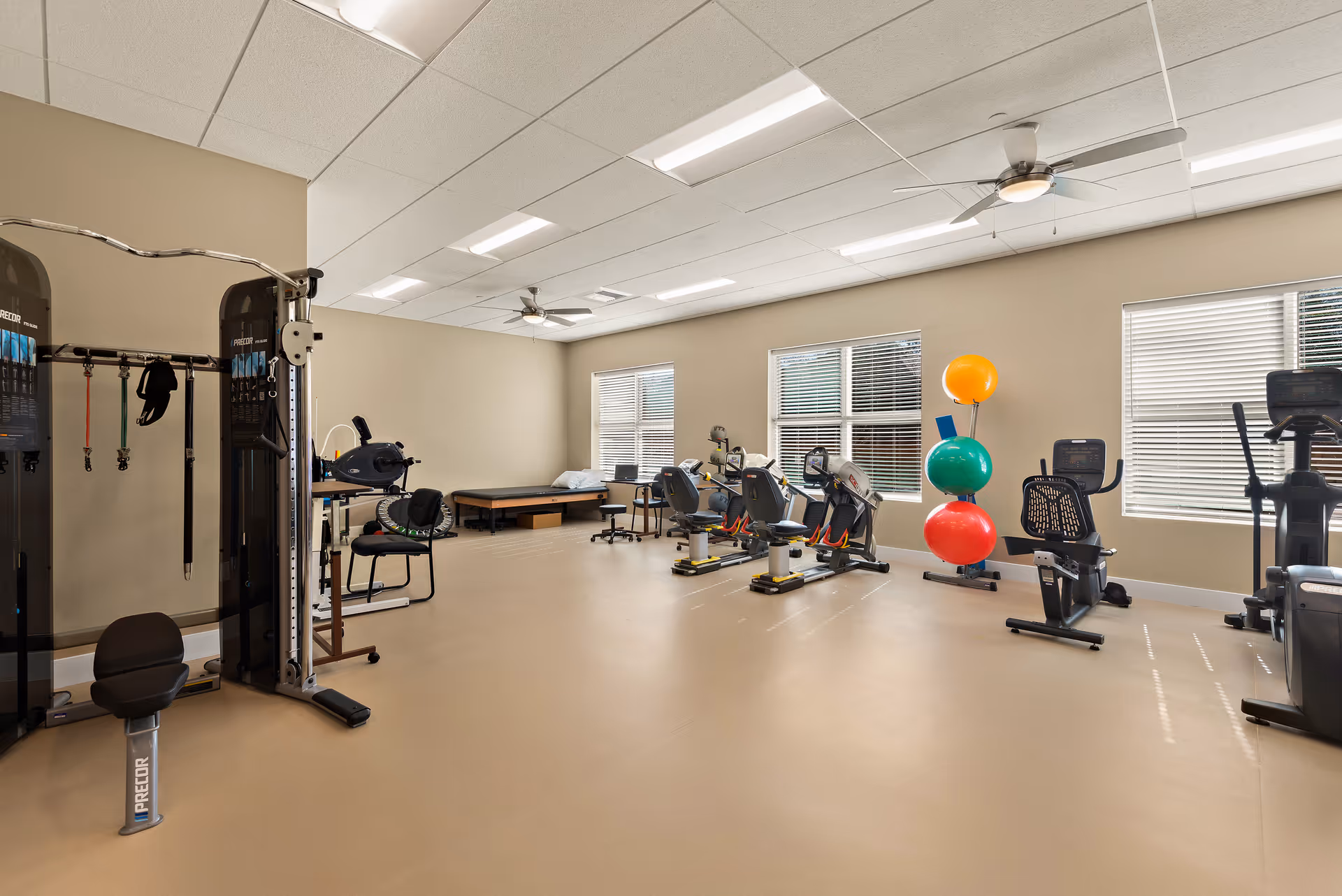 A spacious fitness room with exercise equipment including stationary bikes, a multi-functional weight machine, and large colorful exercise balls stacked near the windows. The room has beige walls, large windows with blinds, ceiling fans, and bright overhead lighting.