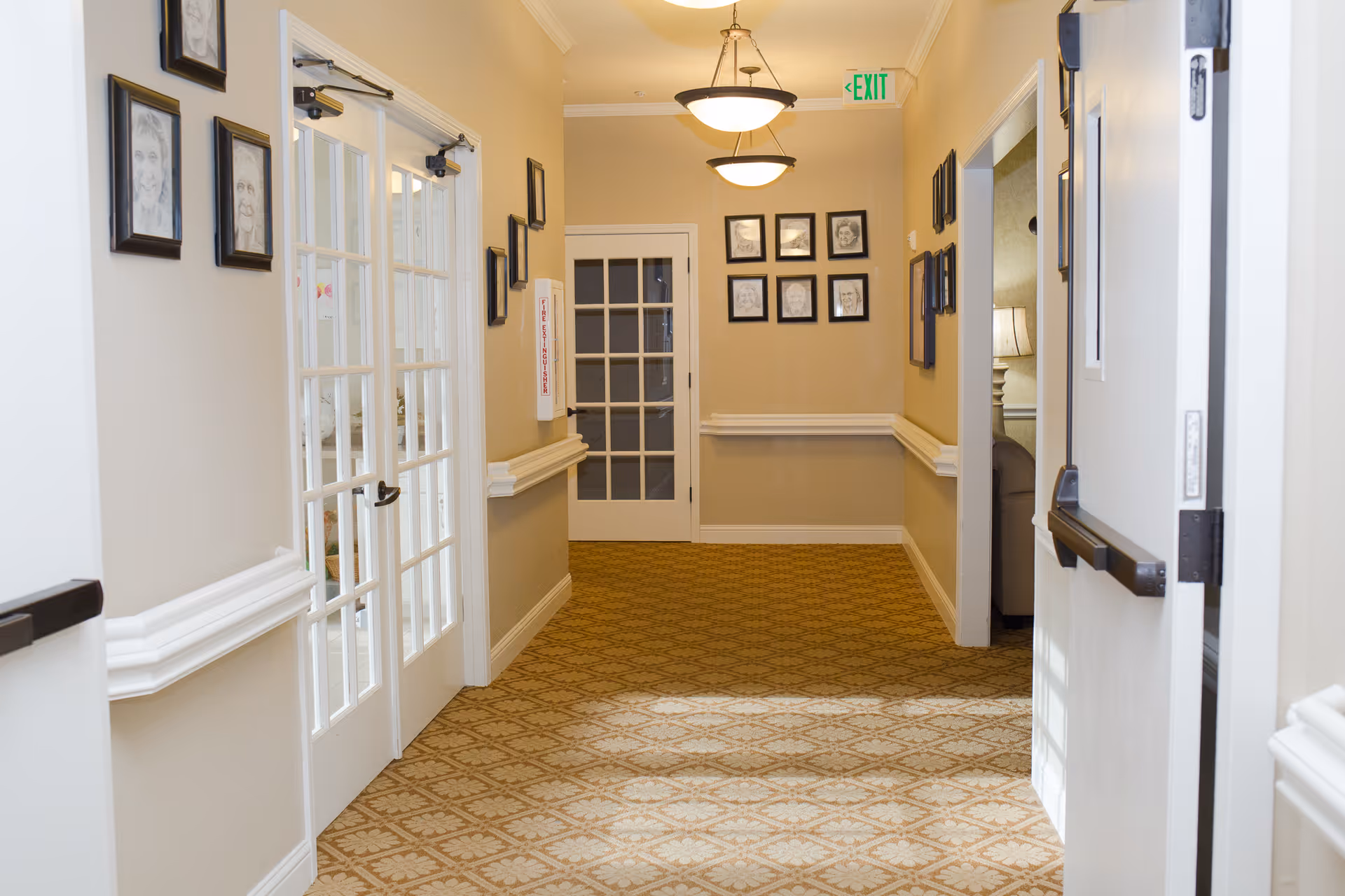 A well-lit hallway in a senior living facility with beige walls, patterned carpet, and white trim. The hallway features framed black-and-white portraits on the walls, two hanging ceiling lights, a white door with glass panes at the end, and double glass doors on the left side. There is an exit sign above the door at the end of the hallway.