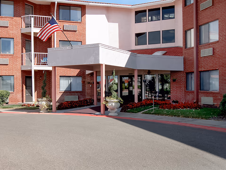 Front entrance of a brick senior living building with a covered porte-cochere, American flag, planters, and flowerbeds.