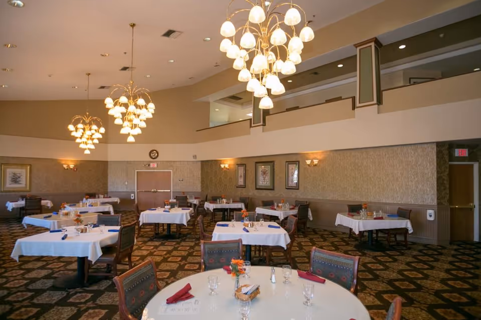 A spacious dining room with multiple tables covered in white tablecloths, each set with glasses, napkins, and small flower arrangements. The room features patterned carpet, beige walls with framed artwork, and several ornate chandeliers hanging from the ceiling. There is a balcony area above and exit doors visible in the background.