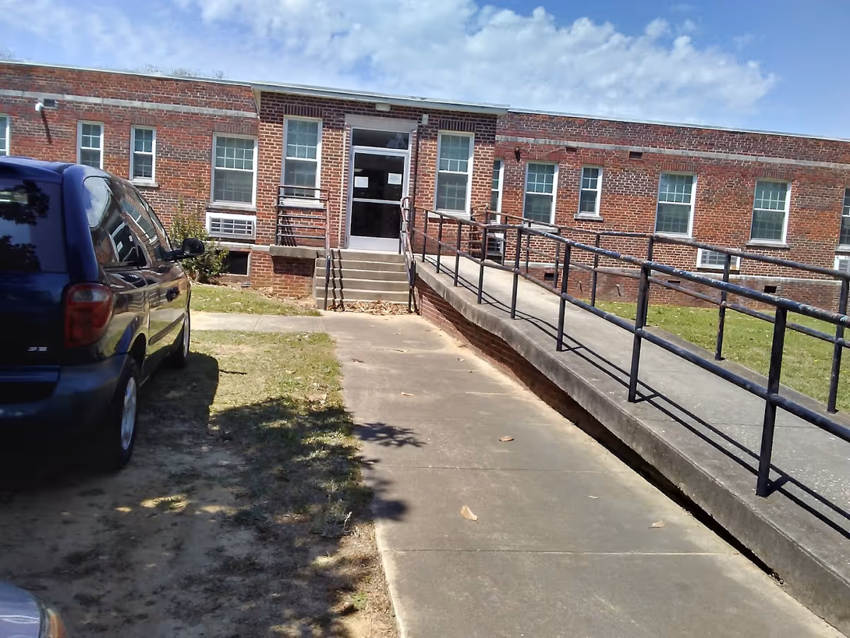 Brick single-story building entrance with a concrete wheelchair ramp, steps, and a parked car nearby.