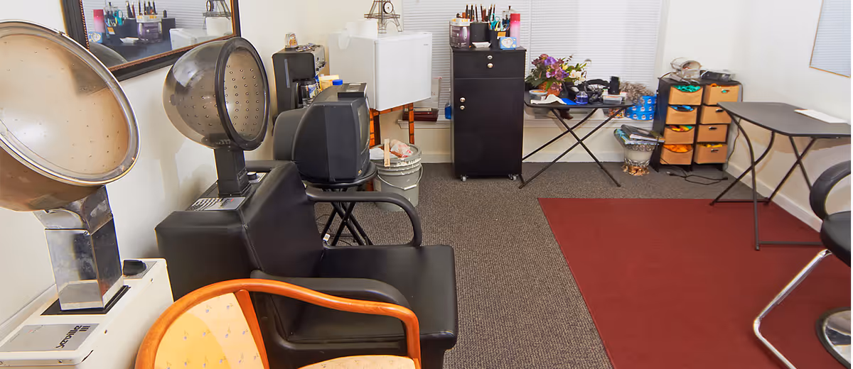 Small salon-style room with hooded hair dryers, black styling chairs, storage cabinets and a red area rug.