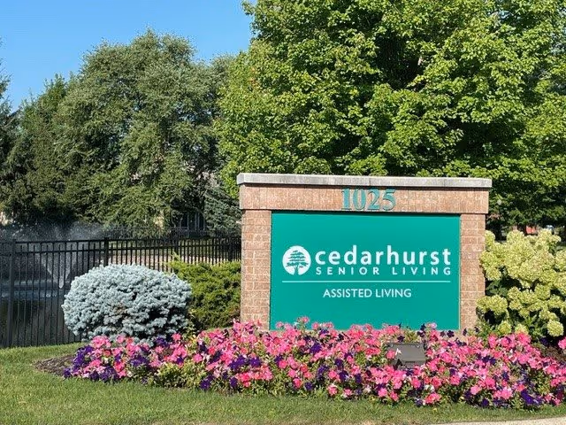 Outdoor view of a brick sign for Cedarhurst Senior Living Assisted Living at 1025, surrounded by colorful flowers and green trees in the background.
