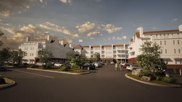 Exterior view of a large, multi-story senior living facility with white walls and brown roofing. The building surrounds a parking lot with several cars parked and a few trees and shrubs planted around the area. The sky is partly cloudy with a warm light suggesting either early morning or late afternoon.