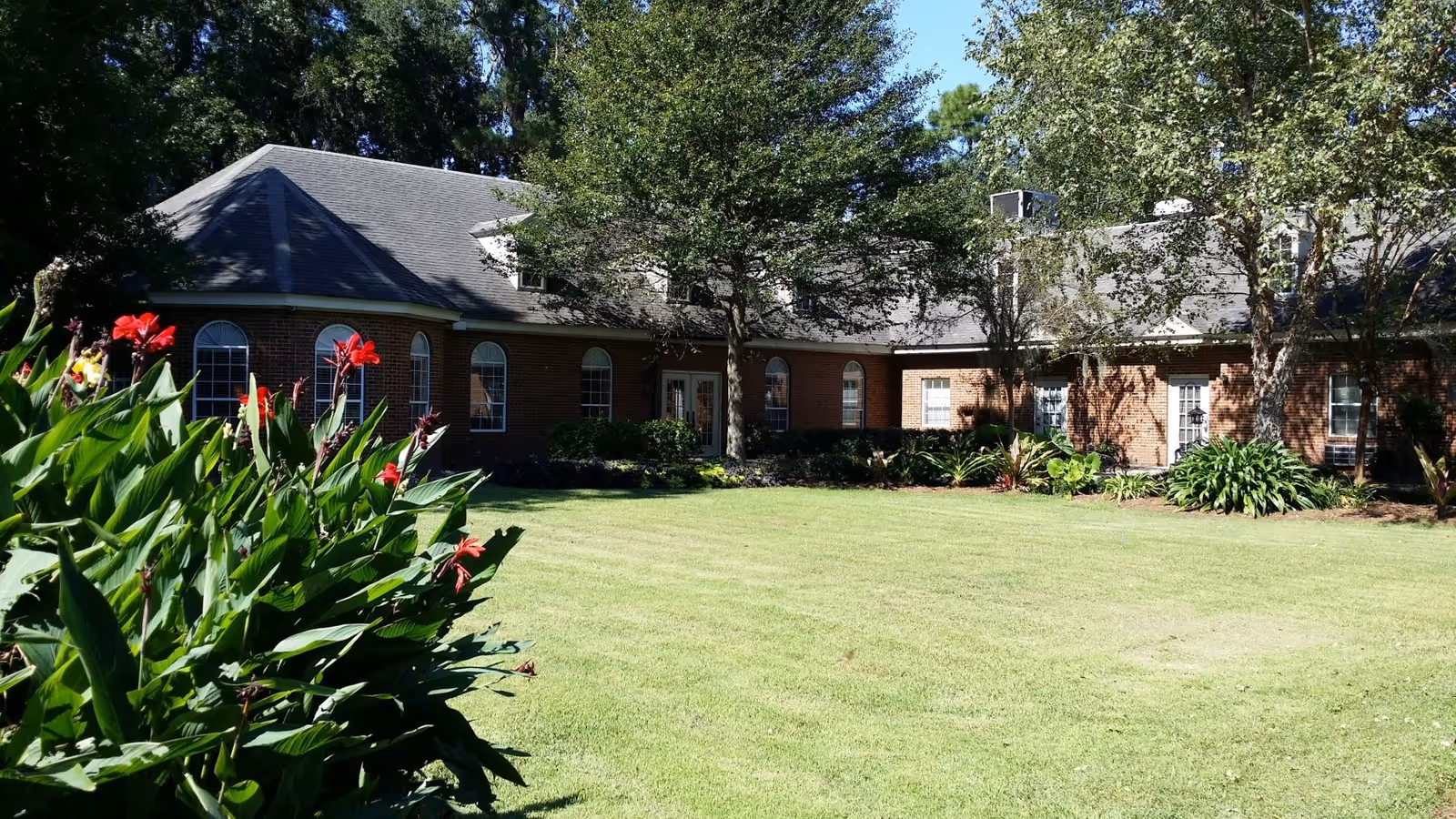 Brick senior living building with arched windows set behind a manicured lawn and flowering plants.