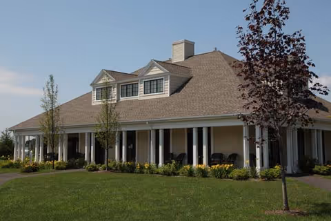 Exterior view of a single-story building with a large covered porch supported by white columns, surrounded by green grass, small trees, and shrubs under a clear blue sky.