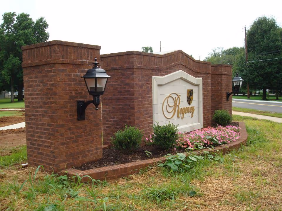 A brick entrance sign for Regency Retirement Village of Morristown with two black lantern-style lights mounted on the brick pillars. The sign is surrounded by a flower bed with pink flowers and green shrubs, located next to a road with trees in the background.