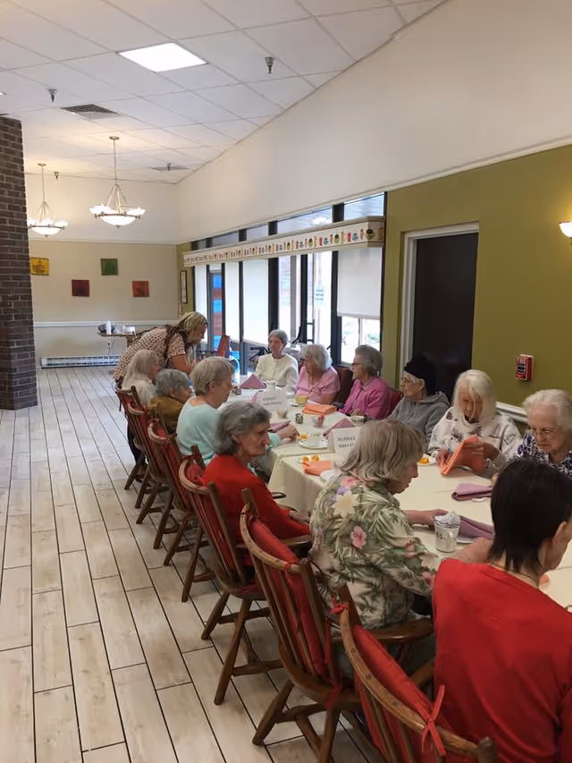 A group of elderly people seated around a long dining table in a well-lit room with large windows. They appear to be engaged in conversation or activities, with a caregiver standing and interacting with them. The room has tiled flooring, green and beige walls, and decorative light fixtures hanging from the ceiling.