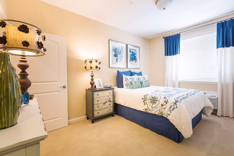 A bright and cozy bedroom with a neatly made bed featuring white and blue patterned bedding. The room has beige walls, a window with white blinds and blue and white curtains, a small nightstand with a decorative lamp and picture frame, and a dresser with a green vase and another lamp with a floral design on the lampshade.