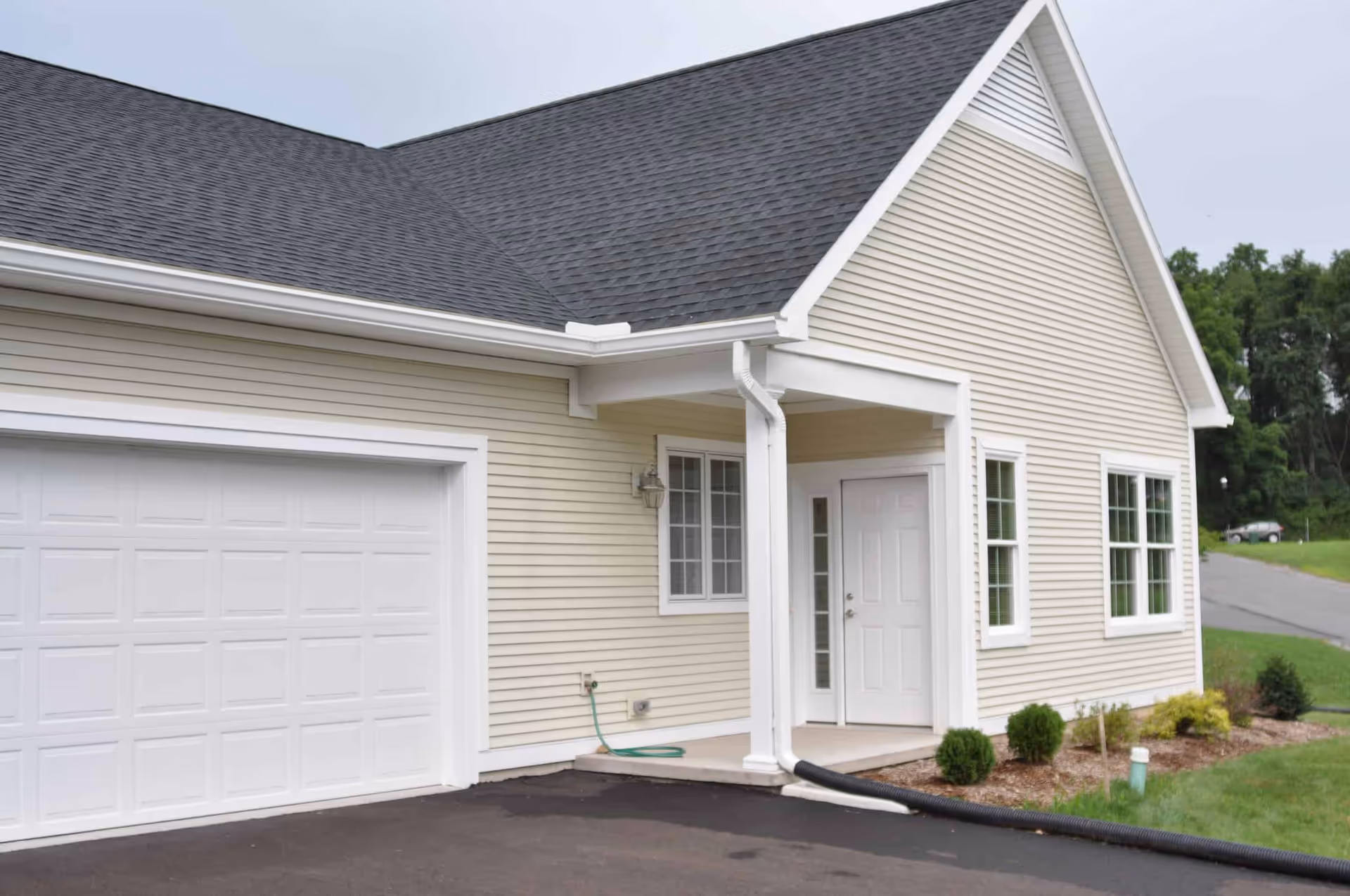 Exterior view of a beige single-story building with white trim, a white garage door, a covered entrance with a white door, and small shrubs planted along the side. The building is surrounded by a paved driveway and green grass with trees in the background.