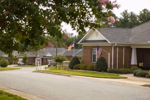 Brick senior living facility with American flags, trimmed shrubs, and a curved driveway.