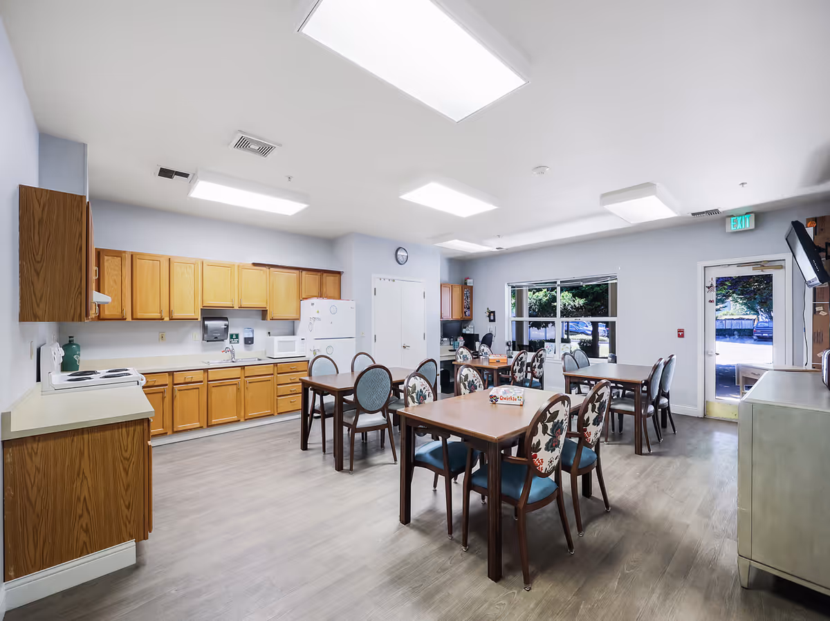 A bright and spacious dining area with several wooden tables and chairs featuring floral upholstery. The room includes a kitchen area with wooden cabinets, a white refrigerator, microwave, and stove. Large windows and a glass door provide natural light and a view of the outside.