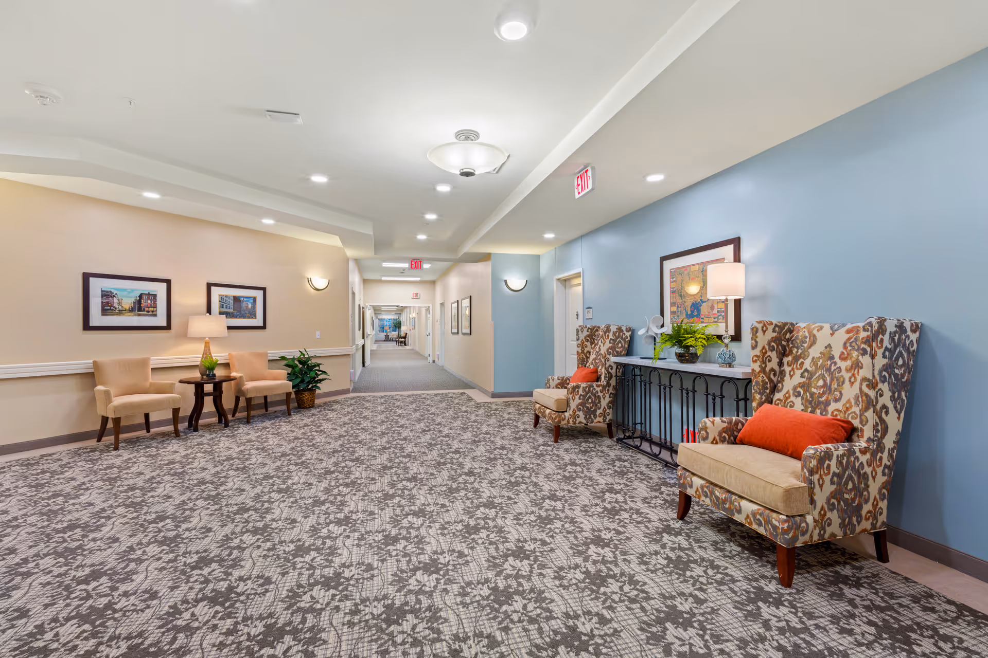 A well-lit hallway in a senior living facility with patterned carpet flooring, beige and blue walls, and several seating areas. On the left, there are two beige chairs with a small round table and a lamp between them, along with framed pictures on the wall. On the right, two patterned armchairs with orange cushions are placed near a console table with a lamp, plants, and framed artwork above it. The hallway extends into the distance with more doors and exit signs visible.