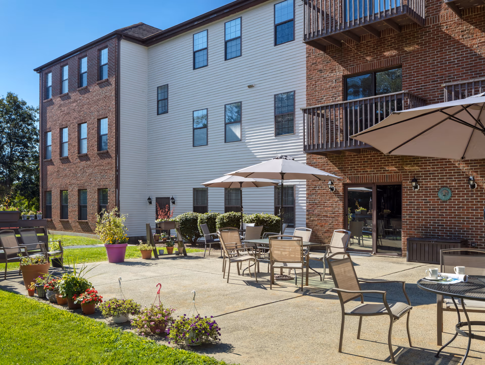 Outdoor patio area at Cedar Woods facility with multiple tables and chairs under large umbrellas, surrounded by potted plants and flowers, adjacent to a brick and white siding building with balconies and windows.