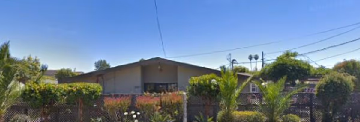 Exterior view of a single-story building with a sloped roof behind a fence with greenery and plants in front. The sky is clear and blue.