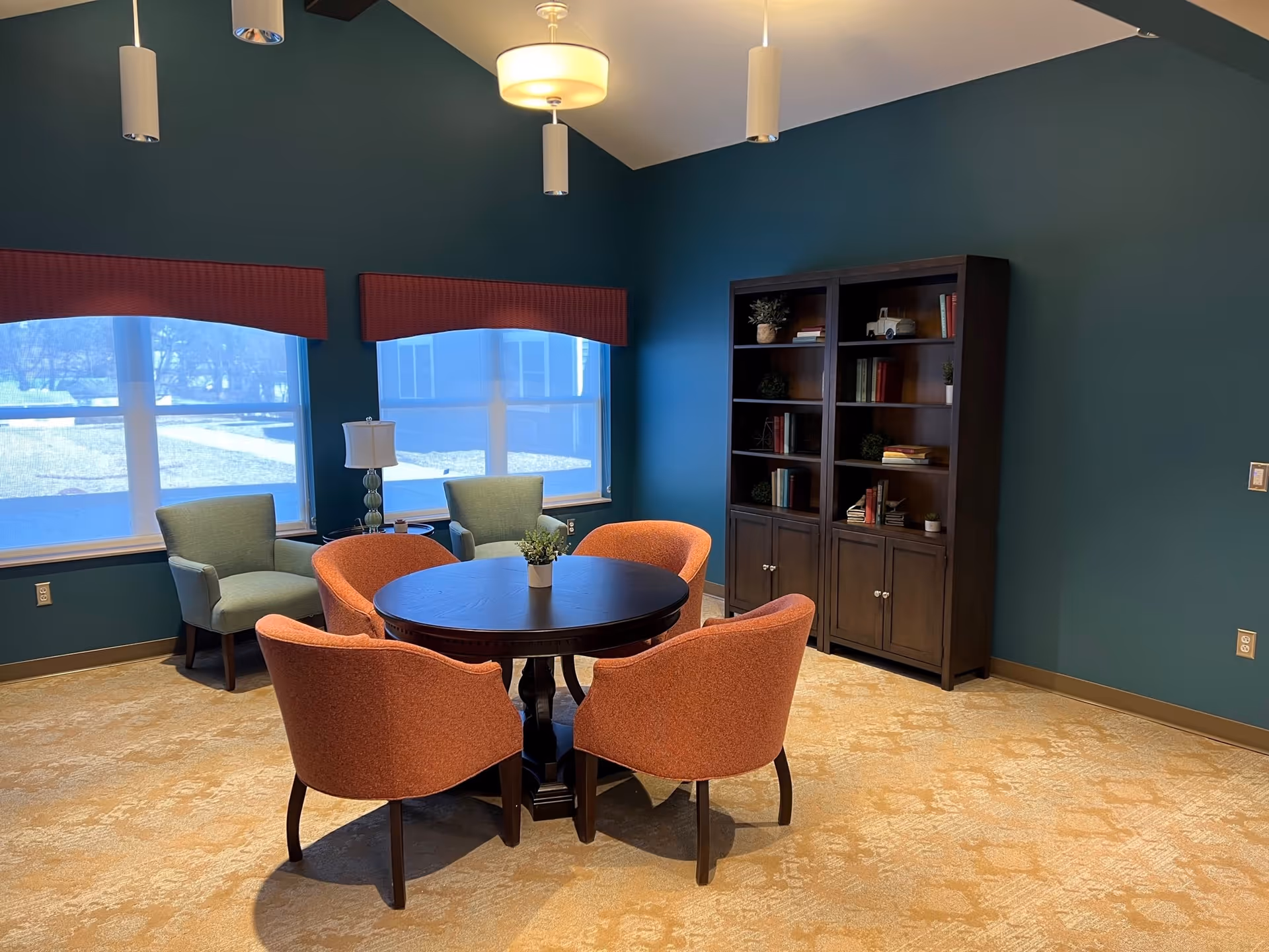 A cozy interior room with teal walls and beige carpet featuring a round wooden table surrounded by four orange upholstered chairs. Two green armchairs are positioned near two large windows with red valances. A dark wooden bookshelf filled with books and decorative items stands against the wall. The room is lit by ceiling pendant lights and a central ceiling light fixture.