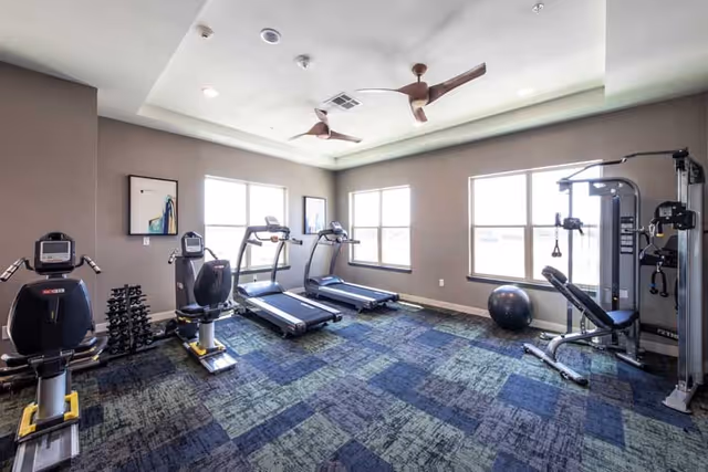 A bright fitness room with two recumbent exercise bikes, two treadmills, a multi-function weight machine, a rack of dumbbells, and an exercise ball. The room has large windows letting in natural light, ceiling fans, and patterned carpet flooring.