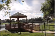 A wooden gazebo with a shingled roof situated on a grassy area next to a body of water, surrounded by a metal fence and trees under a partly cloudy sky.