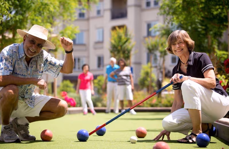 A group of smiling seniors playing lawn bowling on a green lawn in front of a residential building.
