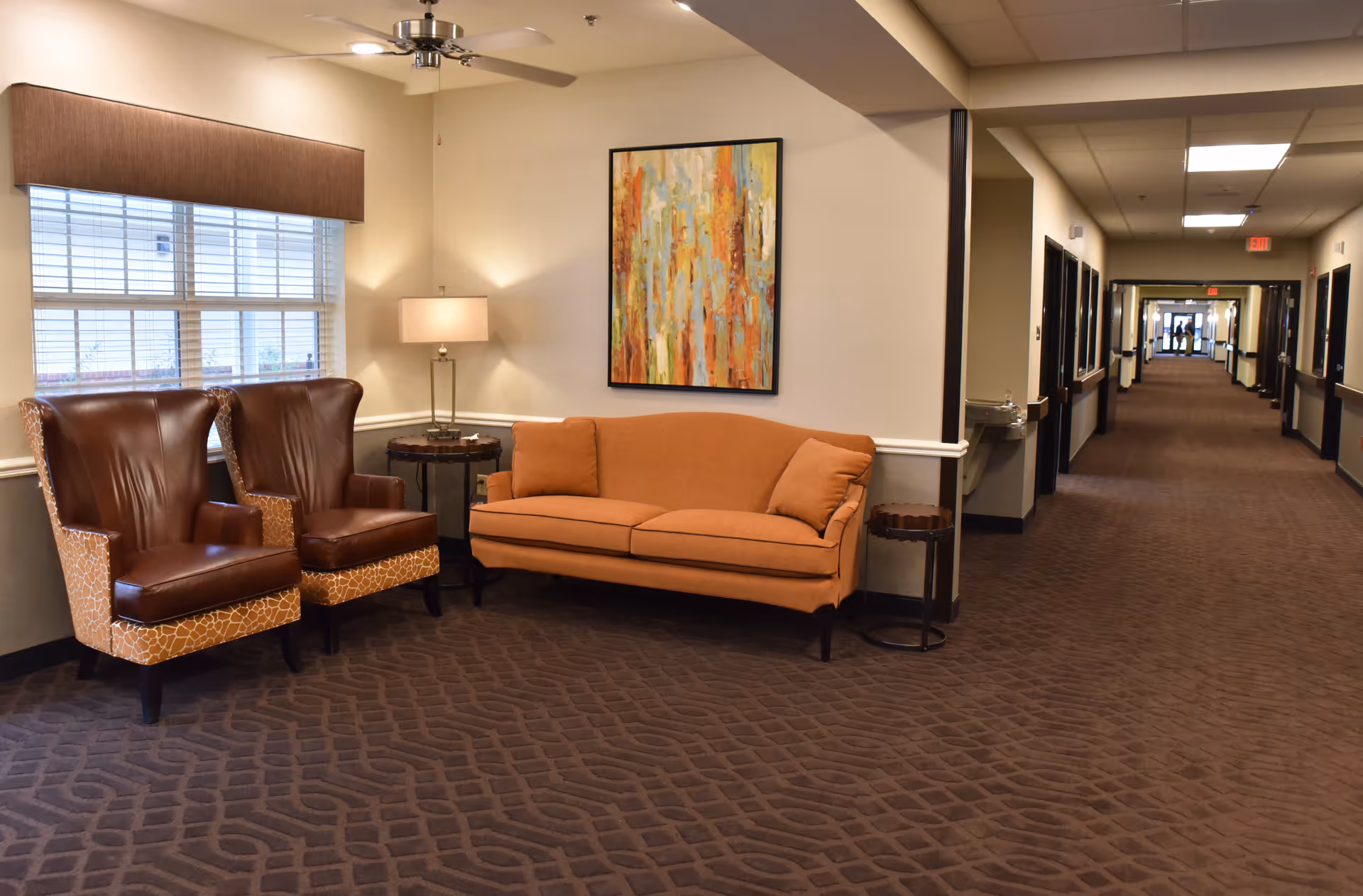 A seating area in a senior living facility hallway with two leather chairs, an orange couch, wall art, and a long carpeted corridor.