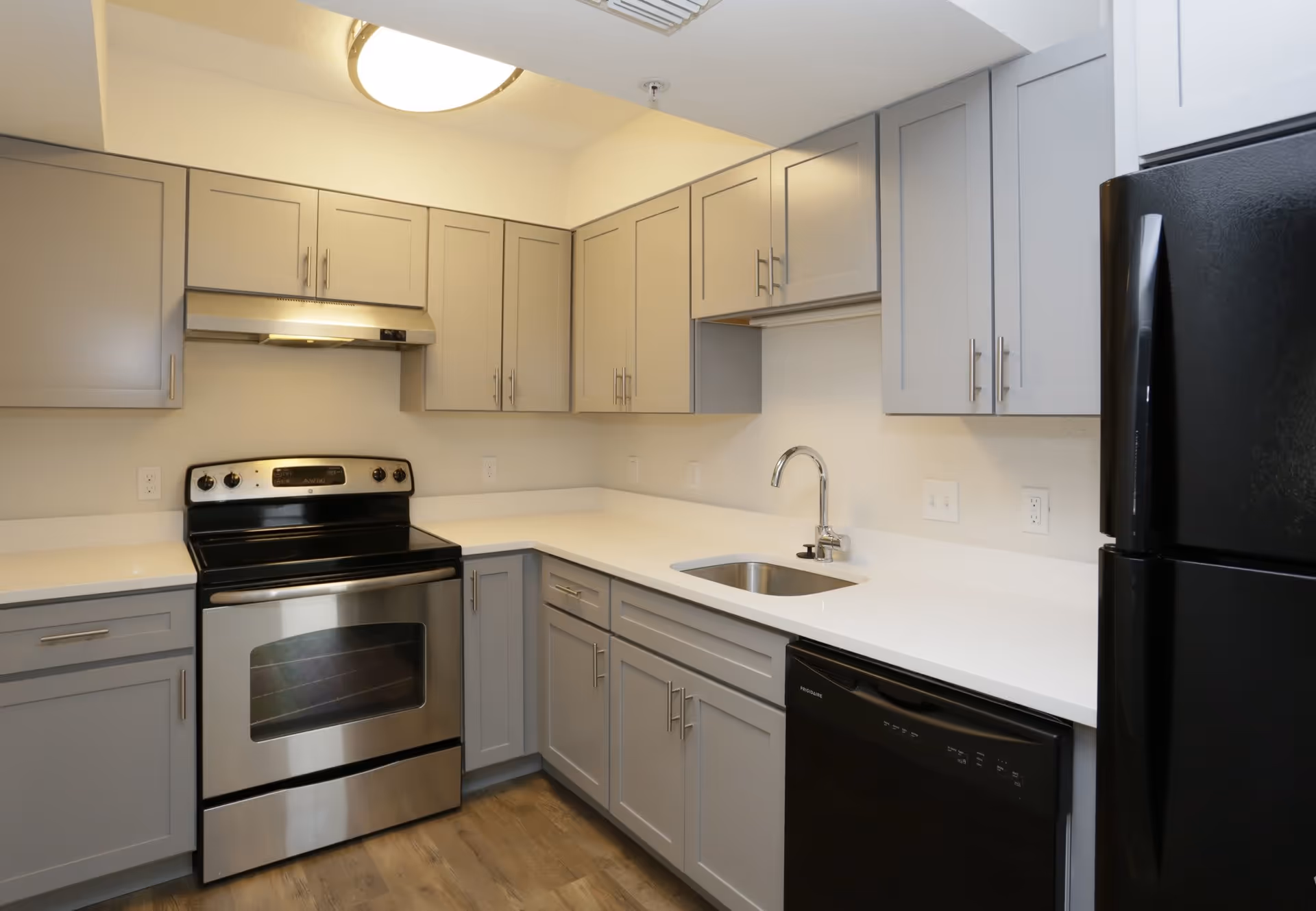 Modern kitchen with light gray cabinets, white countertops, stainless steel oven with range hood, black dishwasher, black refrigerator, and a small stainless steel sink with a chrome faucet. The floor is wood-style and the kitchen is well-lit with a ceiling light.