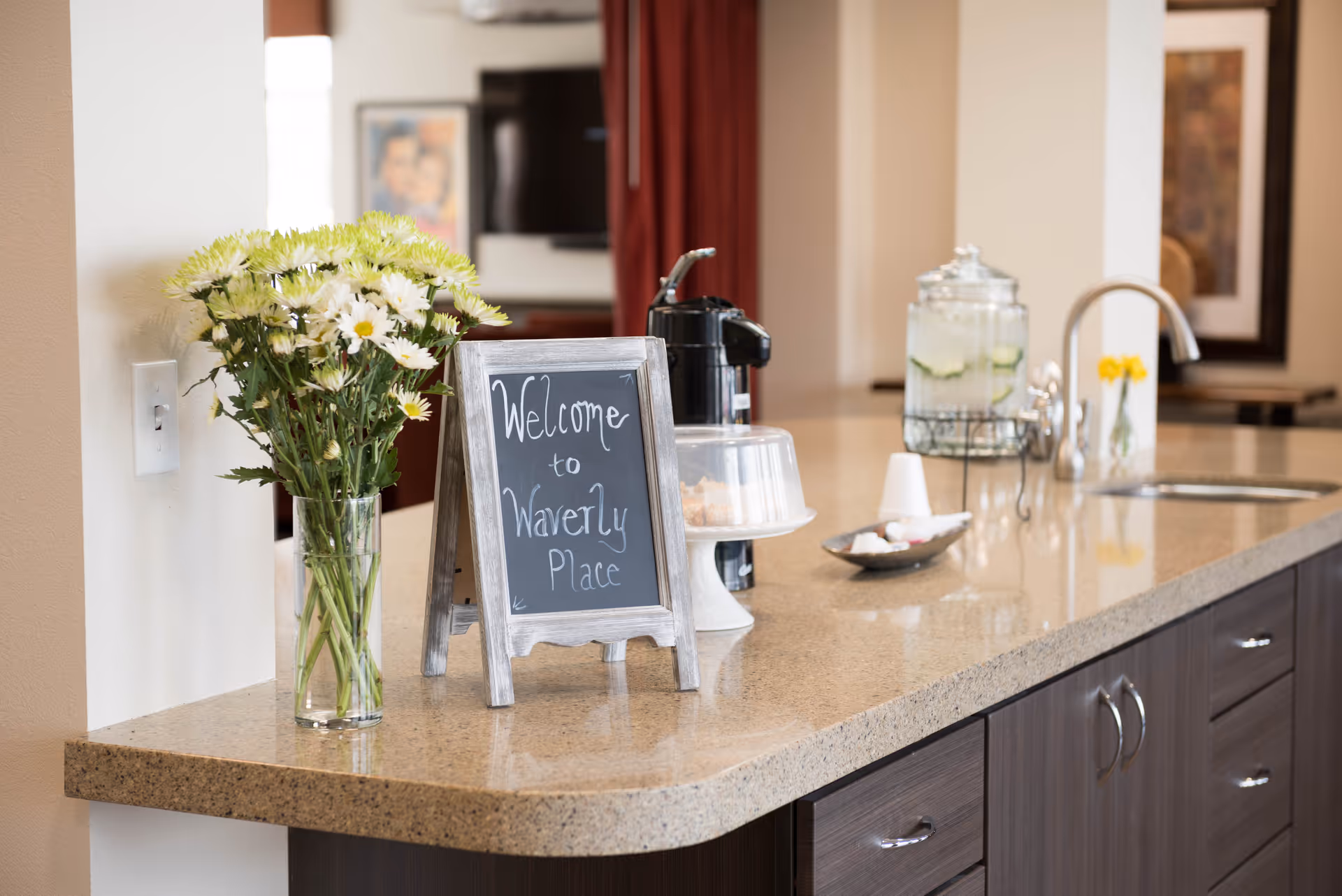 A kitchen countertop with a vase of white and yellow flowers, a small chalkboard sign that reads 'Welcome to Waverly Place', a coffee maker, a covered cake stand, a glass container with infused water, and a sink with a modern faucet.