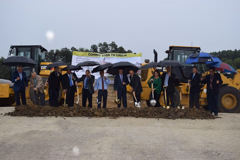 A rainy groundbreaking ceremony with people holding umbrellas and shovels in front of construction equipment and a 'Coming Soon to Green' banner.