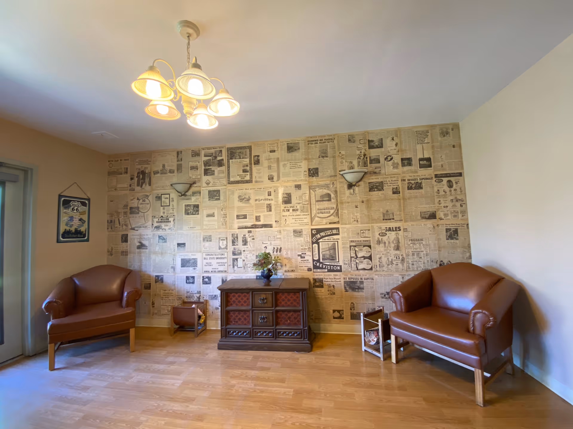 A cozy sitting area with two brown leather armchairs facing each other, a wooden cabinet with decorative drawers in the center, and a wall covered in vintage newspaper clippings. The room has wooden flooring, a ceiling light fixture with five lamps, and a framed Route 66 sign on the left wall near a glass door.