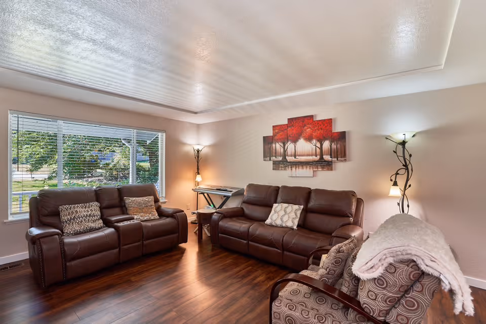 A cozy living room with two brown leather sofas and a patterned armchair with a white throw blanket. There are decorative pillows on the sofas, a multi-panel wall art featuring red trees, a keyboard on a stand in the corner, and two floor lamps providing warm lighting. A large window with blinds shows greenery outside.